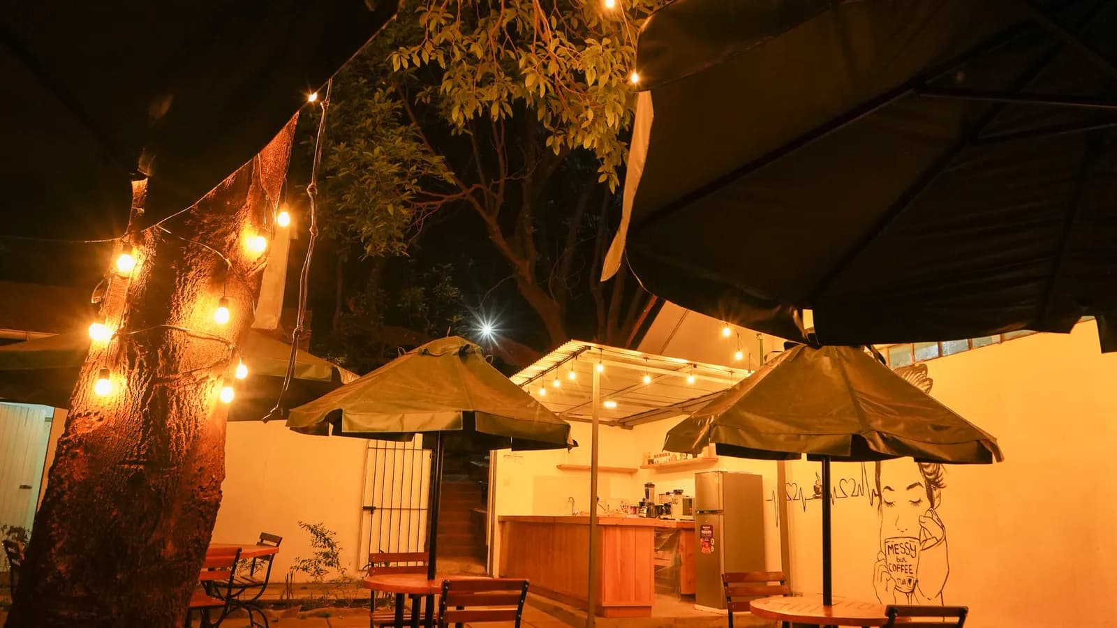 The coffee shop terrace at night, with umbrellas, wooden tables and string lights in a tree